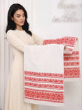 A woman in a cream-colored dress holds up a white long towel or fabric panel featuring red Slavic geometric patterns along the bottom and sleeves, set against a white paneled wall. The long towel is used on wedding for bread and salt ceremony.