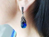 A long earrings made of silver with marcasite stones and royal blue faceted topaz gemstone on a woman earlobe with black hair till the middle of neck.