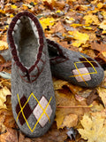 A pair of handmade gray wool felt slipper shoes with argyle patterns in dark brown, light pink, and yellow, displayed on a bed of fallen autumn leaves.