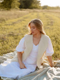 A woman sits on a blanket in a sunlit grassy field, wearing a white dress with a cream-colored sleeveless merino wool vest with zipper and large pockets. 