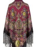 A detailed view of a woman wearing an ornate Slavic-style winter scarf made from 100% merino wool. The heavy, warm fabric is draped closely around her face, highlighting the fine, multicolored paisley scrollwork on the deep red top layer and the large, artistic rose flower prints on the black fabric resting on her shoulder.