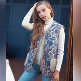 A woman wearing a sleeveless blue and beige floral-patterned sheepskin vest over a white turtleneck and light blue jeans, shown in a room setting. 