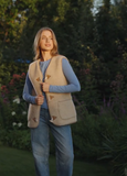 A woman wearing a beige fleece vest with brown patchwork details over a light blue long-sleeve top and blue jeans, standing in a garden setting.