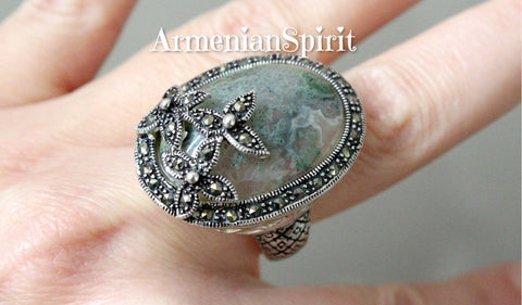 Expertly handcrafted in Armenian design, this sterling silver 925 ring features a large oval moss agate gemstone, accented with marcasite flowers. The big cabochon and vibrant green hue elevate any outfit, making it the perfect statement piece. Enjoy its timeless elegance and superior quality.
