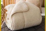 A rolled cream-colored natural merino wool sheepskin blanket is displayed on a bed with a quilted dark gray cover and a wooden headboard in the background. 