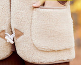Close-up of a beige sherpa fleece jacket with brown toggle closures and a front pocket, worn by a person whose hand is visible inside the pocket.