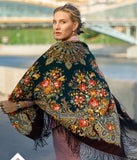 A woman wrapped in an oversized merino scarf adorned with a vintage lily and peony pattern. The harmonious blend of deep green and chocolate  brown tones highlights the retro style, while the soft natural fibers and silky fringe add a touch of luxury.