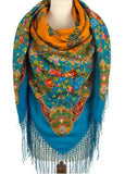 A warm and cozy winter look featuring a maneqeun wrapped in a large, blanket-style merino wool scarf decorated with traditional orange and blue floral motifs.