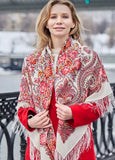 A medium shot of a smiling blonde woman looking to the side, wearing a bright red coat. She is wrapped in a large, oversized ivory merino wool shawl featuring an intricate traditional Slavic design. The fabric is decorated with heavy pink and orange rose patterns, sweeping paisley swirls, and long, delicate white fringes hanging down the front.