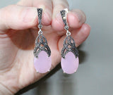 Pink teardrop earrings with silver and black detailing held in a hand against a neutral background