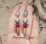 A pair of silver Armenian earrings featuring garnet stones and pomegranate-shaped designs, held in a person's hand.