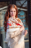 Woman wearing a floral merino wool white shawl with small red roses in front of a store window. A detailed close-up shot of a luxurious Slavic-style merino wool shoulder shawl worn by a ginger woman. The oversized scarf displays a rich botanical border design filled with blooming red vintage roses and complex yellow paisley ornaments, draped to reveal the folds of the thick fabric.