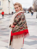 Woman wearing a colorful red patterned merino wool shawl with red fringe on a city street. A street-style photograph of a woman wrapped in a warm, premium merino wool shawl. The scarf features a vibrant, colorful pattern on a deep red base with textured red fringes along the hem, captured in a bright urban city setting.