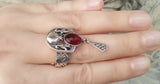 Silver pomegranate ring featuring garnet stones and pomegranate-shaped designs, held in a person's hand.