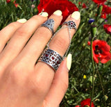 A person's hand showcasing two sterling silver rings, one with a double design and the other a knuckle ring, with no visible red stones. The hand is set against a background with red floral elements.