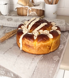 A round decorated sweet bread loaf with piped cream-colored rose and wheat designs on top, resting on a patterned beige Slavic table runner in a kitchen setting.
