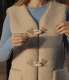 A close-up of a person wearing a beige sherpa fleece sleeveless vest with tan toggle closures over a light blue long-sleeve shirt.