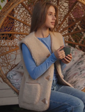 A woman sits in a woven hanging chair wearing a beige modern woolen sleeveless toggle borg vest over a blue long-sleeve top and blue jeans, holding a dark green mug.