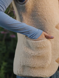 Close-up of a person wearing a cream-colored sherpa fleece teddy woolen vest with a large front patch pocket, shown outdoors with a blue long-sleeve shirt.