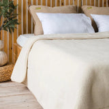 A cozy bedroom scene showing a neatly made bed with a cream-colored natural merino wool fur blanket cover, white pillows, and tan accent pillows in front of a light wood slat headboard. 