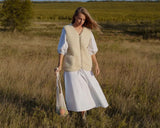 A woman stands in a grassy outdoor field wearing a cream-colored merino wool vest over a white dress, holding a woven tote bag. 