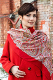 Woman wearing a red coat and patterned large red shawl with pale yellow and green ornament against a brick wall.