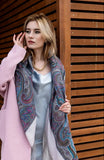 Woman wearing a pink coat and gray patterned merino wool scarf against a wooden background. A studio mannequin showcases a cozy winter blanket shawl made from 100% merino wool. The design blends heritage Slavic patterns in soft ivory, muted gray, and slate blue, draped heavily to show off its thick texture and flowing fringe.