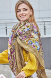 Woman wearing a colorful patterned scarf. The shawl has a yellow base with pale blue and purple flowers. The material is high quality merino wool and silky fringe.