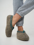 A person wearing gray knit pants and felt slip-on slippers in a warm taupe-brown color with teal blue trim, shown against a light gray studio background.