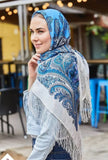 Woman wearing a blue and white patterned scarf with fringe, standing outdoors. A milky white floral shawl with light blue flowers in a square shape, calm and refined.