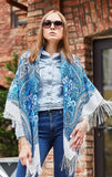 Woman wearing a blue and white patterned shawl over a denim outfit against a brick wall. This milky white square Slavic fringed shawl features light blue floral patterns for a gentle, classic style.