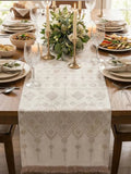 A beige patterned Slavic traditional table runner with fringe is styled on a wooden dining table set for a meal, featuring candles, wine glasses, plates, and a floral centerpiece. 