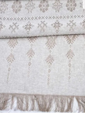 A folded light beige woven linen table runner with a traditional geometric pattern, featuring fringed edges.