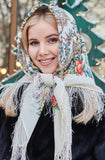 A festive portrait of a woman elegantly showcasing a traditional ivory woolen shawl. The blanket-style merino wrap serves as a head covering, presenting a vivid contrast between the neutral white fabric, the colorful orange-red floral bouquets, and her dark coat.