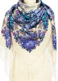 A manequin wears a large, square-shaped merino wool ivory scarf draped over her shoulders. The fabric has a beige base covered in a detailed pattern of bright blue and soft purple rose flowers, complete with textured fringes along the hem.