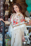 Woman wearing a floral merino wool white shawl in a store setting. A woman models a warm, blanket-style winter fringed Slavic shawl made from 100% merino wool. The opaque white fabric features a striking contrast with its heavy, multicolored folk pattern of red, yellow, and blue flowers, conveying a very cozy and classic Slavic cultural aesthetic.