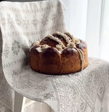 A large braided loaf of bread, likely a Slavic or similar sweet bread, decorated with cream-colored icing, sitting on a chair draped with a patterned beige long traditional table runner with Slavic ornaments.