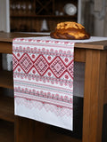 A white Slavic table runner with red and dark gray traditional geometric patterns draped over a wooden table, with a loaf of bread placed on top.