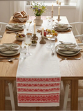 A white table runner with a red Slavic folk pattern is displayed on a wooden dining table set for a meal.