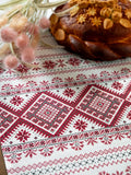 A close-up of a white embroidered Slavic tablecloth with traditional red and gray geometric patterns, styled with a decorated brown bread loaf and soft pink dried flowers in the background.