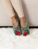 A person wearing gray felted slip-on slippers with red berry and green leaf embroidery, standing on a white faux-fur rug in front of a light beige wall.