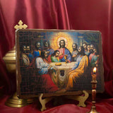A rectangular religious icon plaque depicting Jesus and the twelve apostles, displayed on a small wooden stand against a draped red satin backdrop, with a brass cross and candle holder nearby.
