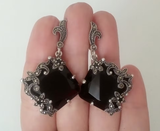A black onyx square earrings with silver 925 marcasite floral details, displayed on a hand.