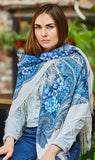 Woman wearing a blue floral patterned scarf with fringe, sitting outdoors. A square shawl in milky white with light blue flowers. Soft colors and a clean, elegant look.