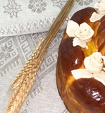 A close-up of a braided bread loaf with glossy browned tops and white rose-shaped frosting decorations, placed on a light gray patterned table cloth next to two wheat stalks.