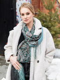 Woman wearing a white coat and patterned scarf in an urban setting. A large green scarf decorated with teal and black paisley patterns. The traditional ethnic style feels classic and refined, adding quiet elegance to any outfit.