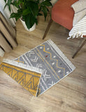 Two patterned gray rugs on a wooden floor with a plant and furniture in the background.