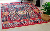 Multicolored geometric Armenian patterned rug in red and blue  on a wooden floor with a plant in the corner.