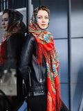 Woman wearing a colorful Slavic floral  headscarf with a black leather jacket, standing against a reflective surface. The base is red and the flowera are light blue and yellow.