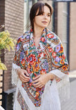 A woman wrapped in an extra-large white merino scarf featuring a traditional Slavic herbal pattern in red and yellow tones. The retro-inspired design is enriched by natural merino softness and finished with a flowing silky fringe.